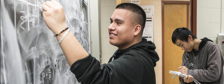 A student does math on a chalkboard while another student writes in a notebook.